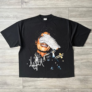 Pode incluir: Uma t-shirt preta de manga curta com uma impressão gráfica de uma pessoa sorridente a usar uma luva de lantejoulas sobre os olhos. O design inclui uma assinatura branca estilizada no canto inferior esquerdo e uma pequena figura a dançar.