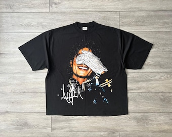 May include: A black short-sleeve t-shirt featuring a graphic print of a smiling person wearing a sequined glove over their eyes. The design includes a stylized white signature at the bottom left and a small figure in a dance pose on the right side.