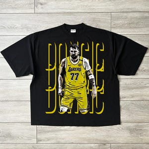 May include: Black t-shirt featuring a graphic of a basketball player in a yellow Lakers jersey with the number 77. The player's name is written in yellow stylized text. The shirt is on a light wood surface.