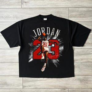 May include: Black t-shirt with a graphic of Michael Jordan in action, the word "JORDAN" above and the number "23" in red. Includes a signature. A classic basketball-themed garment.