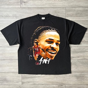 以下が含まれることがあります： 黒の半袖Tシャツで、バスケットボール選手の顔の大きなグラフィックプリントと、赤いジャージを着た同じ選手の全身の小さな画像が特徴です。選手の顔の下には1.NTという文字が白でプリントされています。