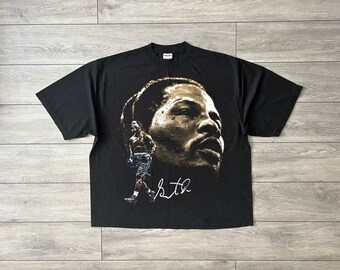 May include: Black t-shirt featuring a sepia-toned portrait of a man's face and a smaller image of a boxer. The shirt also includes a signature in white. The shirt is made of a soft material.