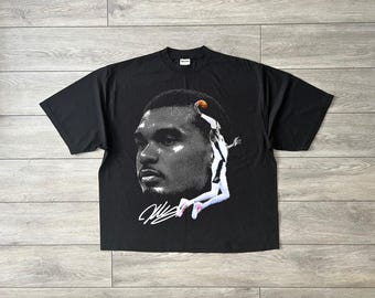 May include: Black t-shirt featuring a grayscale image of a basketball player mid-air, with a basketball in hand. The design includes a close-up of the player's face and a signature in white. The shirt is made of a soft material.