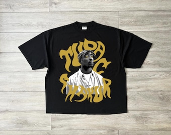 May include: Black t-shirt featuring a grayscale image of a man with a bandana and gold text that reads "TUPAC SHAKUR". The graphic is in a stylized font. The shirt has short sleeves and a crew neck.