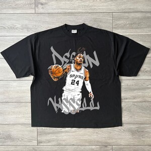 Pode incluir: Camiseta preta com um gráfico de um jogador de basquete segurando uma bola de basquete. O jogador está usando um uniforme branco e preto com o número 24. O nome "Dejounte Murray" está escrito em estilo grafite.