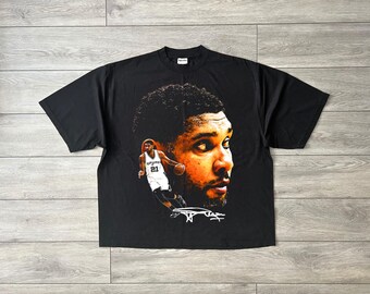 May include: A black short-sleeve t-shirt featuring a large graphic print of a basketball player's face in warm tones, a smaller full-body image of the same player in a white jersey with the number 21, and a white signature graphic at the bottom.