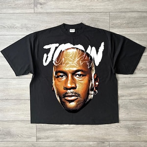 May include: Black t-shirt with a portrait of Michael Jordan and the name "JORDAN" in white above the image. The graphic is a detailed illustration of Jordan's face. The shirt is made of a soft material.