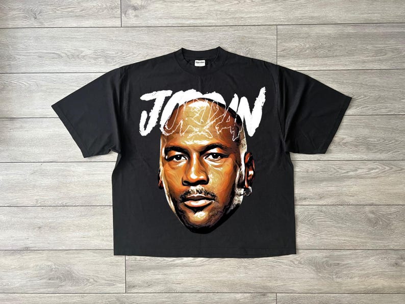 May include: Black t-shirt with a portrait of Michael Jordan and the name "JORDAN" in white above the image. The graphic is a detailed illustration of Jordan's face. The shirt is made of a soft material.