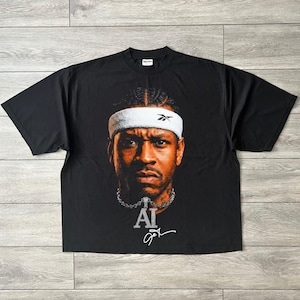 May include: A black short-sleeve t-shirt featuring a large graphic print of Allen Iverson wearing a white Reebok headband. Below the portrait is a silver chain necklace with an AI pendant and a white signature graphic printed on the chest.