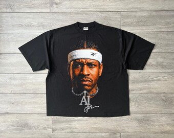 May include: A black short-sleeve t-shirt featuring a large graphic print of Allen Iverson wearing a white Reebok headband. Below the portrait is a silver chain necklace with an AI pendant and a white signature graphic printed on the chest.