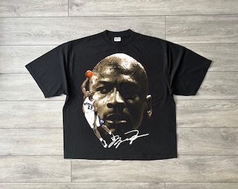 May include: A black short-sleeve t-shirt featuring a large graphic print of Michael Jordan's face in a sepia tone. Below the face is a smaller image of Jordan in a white jersey dunking a basketball, accompanied by a white signature graphic.