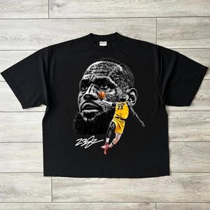 May include: Black t-shirt featuring a graphic of a basketball player's face and a player in a yellow uniform with the number 23. The design includes a signature in white. The shirt is made of a soft material.