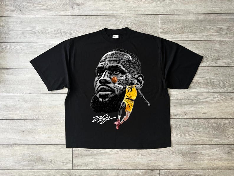 May include: Black t-shirt featuring a graphic of a basketball player's face and a player in a yellow uniform with the number 23. The design includes a signature in white. The shirt is made of a soft material.