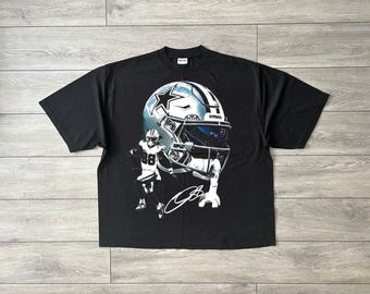 May include: A black short-sleeve t-shirt featuring a large graphic print of a silver Dallas Cowboys football helmet, a football player in uniform with the number 88, and a white signature. The design is centered on the front of the shirt.