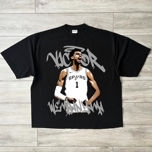 Könnte beinhalten: Schwarzes T-Shirt mit einer Grafik eines Basketballspielers in einem weißen Trikot mit der Nummer 1 und dem Wort "SPURS". Der Name des Spielers "Victor" ist in grauen Graffiti-Buchstaben über dem Spieler geschrieben.