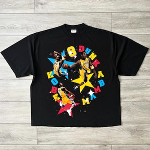 May include: Black t-shirt featuring a colorful graphic of a basketball player in action, with the words "8 Dunk Mamba Kobe" in a starburst design. The player is wearing a yellow and purple uniform. The shirt is made of a soft material.