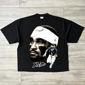 Puede incluir: Camiseta negra con un gran gráfico de la cara de un jugador de fútbol americano con una cinta blanca en la cabeza y el logotipo de los Philadelphia Eagles. También hay una imagen más pequeña del jugador con el uniforme y una firma.