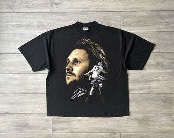 May include: A black short-sleeve t-shirt featuring a large graphic print of a man's face, a football player in a white jersey with the number 17, and a white signature. The design is centered on the front of the shirt against a solid black background.