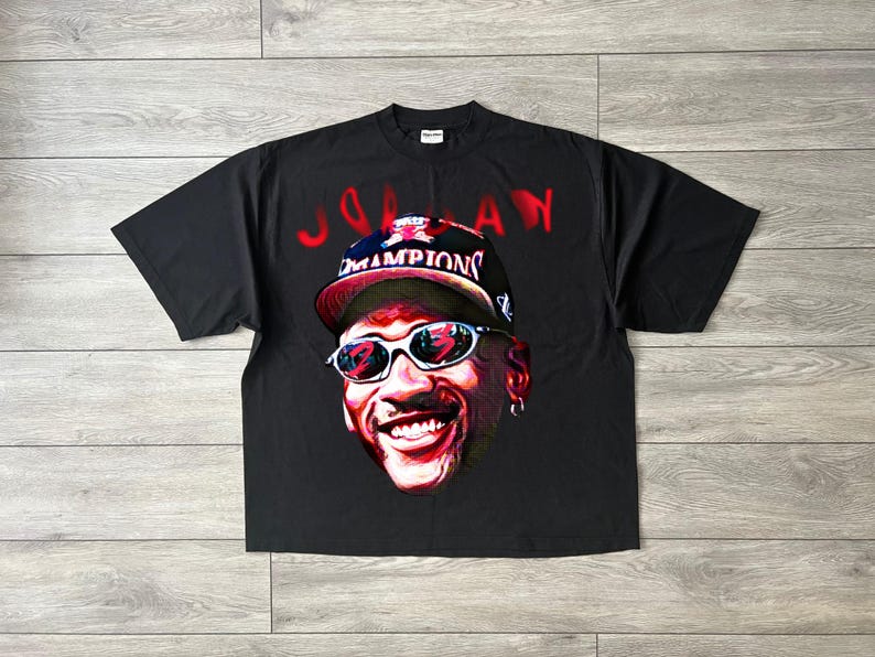 May include: Black t-shirt with a colourful portrait of a smiling person wearing sunglasses and a baseball cap with the word "Champions". The word "Jordan" is written in red above the portrait.