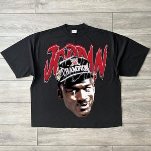 May include: Black t-shirt with a graphic of Michael Jordan wearing a Champion hat. The word "JORDAN" is in red above the image. The shirt has a crew neck.
