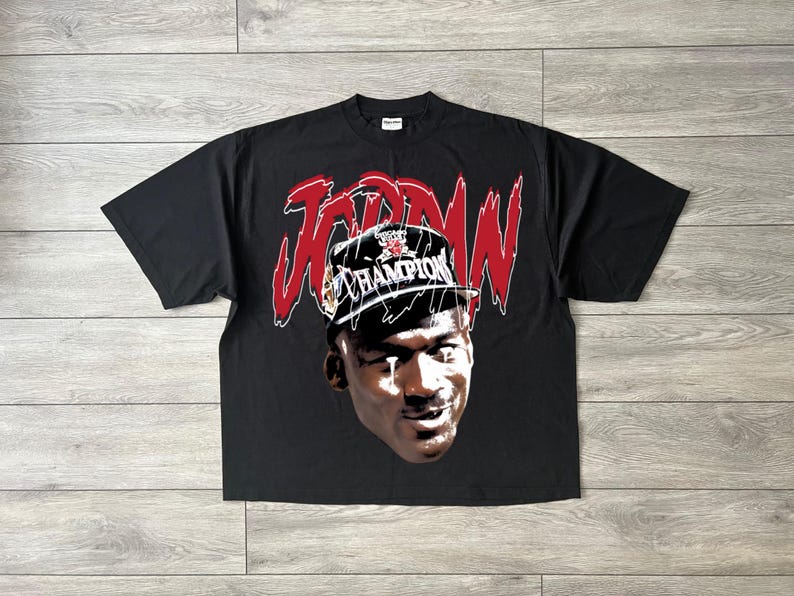 May include: Black t-shirt with a graphic of Michael Jordan wearing a Champion hat. The word "JORDAN" is in red above the image. The shirt has a crew neck.