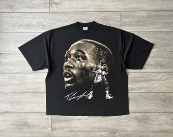 May include: Black t-shirt featuring a sepia-toned portrait of a boxer's face and a smaller image of the boxer in action. The shirt also has the boxer's signature in white. The shirt is made of a soft material.