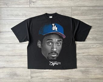 May include: Black t-shirt featuring a grayscale portrait of a man wearing a blue baseball cap with the letters "LA". The shirt also includes a signature in white. The shirt is laid flat on a wooden surface.