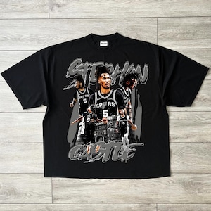 May include: Black t-shirt featuring a graphic of a basketball player in a Spurs jersey. The design includes the player's name, "STEPHAN CASTLE," in a stylized font. The shirt has a crew neck and short sleeves.