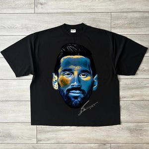 Puede incluir: Camiseta negra con un retrato estilizado de un hombre con acentos azules y amarillos, posiblemente un deportista. El dise&ntilde;o incluye una firma. La camiseta est&aacute; colocada sobre una superficie de madera.