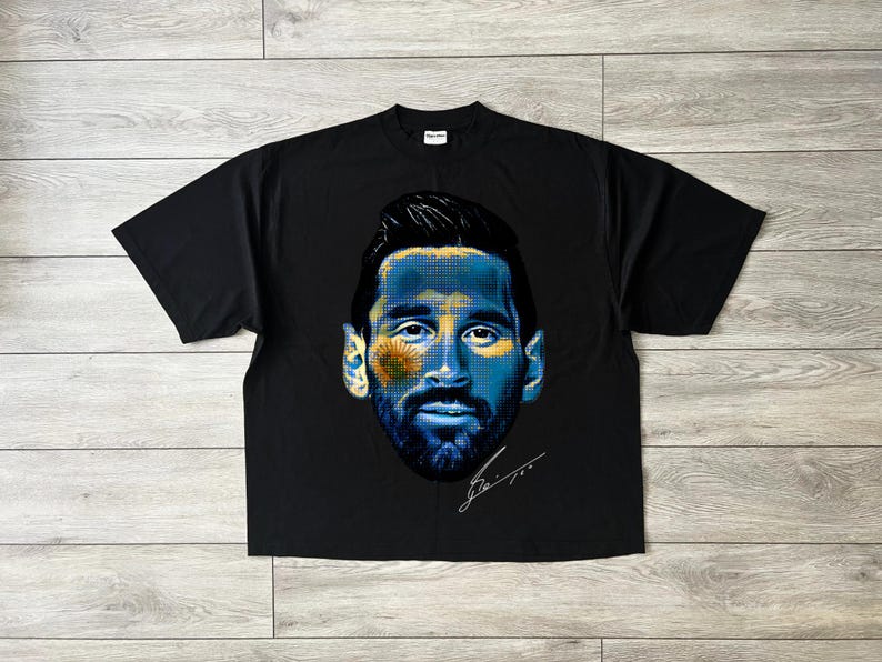 Puede incluir: Camiseta negra con un retrato estilizado de un hombre con acentos azules y amarillos, posiblemente un deportista. El dise&ntilde;o incluye una firma. La camiseta est&aacute; colocada sobre una superficie de madera.