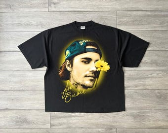 May include: A black short-sleeve t-shirt featuring a graphic print of Justin Bieber wearing a green baseball cap. A yellow flower is positioned near the face, surrounded by a yellow glow. The name Justin Bieber is written in yellow cursive script.