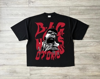 May include: Black t-shirt featuring a grayscale image of a man wearing sunglasses and a hat. The words "B.I.G. Notorious" are printed in red, stylized font across the front of the shirt. The shirt is made of a soft material.
