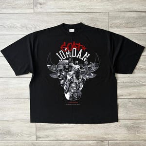 May include: Black t-shirt with a bull's head graphic and the text "GOAT JORDAN" in red and white. The bull's head is filled with images of a basketball player. The shirt is made of a soft material.