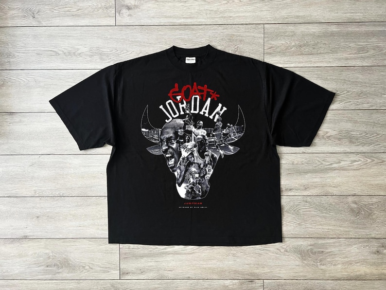 May include: Black t-shirt with a bull's head graphic and the text "GOAT JORDAN" in red and white. The bull's head is filled with images of a basketball player. The shirt is made of a soft material.