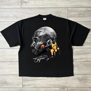 May include: Black t-shirt featuring a grayscale portrait of a basketball player with a basketball and a smaller image of the player in action, wearing a yellow and purple uniform. The shirt includes the player's signature.