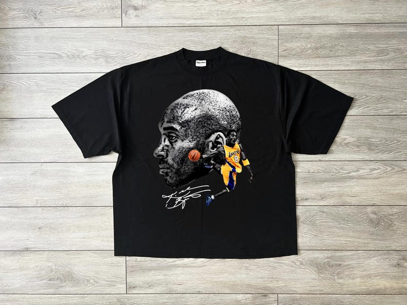 May include: Black t-shirt featuring a grayscale portrait of a basketball player with a basketball and a smaller image of the player in action, wearing a yellow and purple uniform. The shirt includes the player's signature.