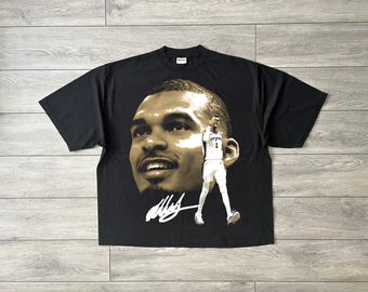 May include: A black short-sleeve t-shirt featuring a large, sepia-toned portrait of a basketball player. To the right of the face is a full-body image of the player in a white uniform with the number 1, and a white signature graphic below.