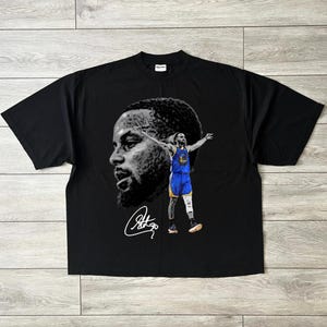 May include: Black t-shirt featuring a grayscale portrait and a full-color image of a basketball player in a blue and gold uniform. The shirt also includes a signature in white.