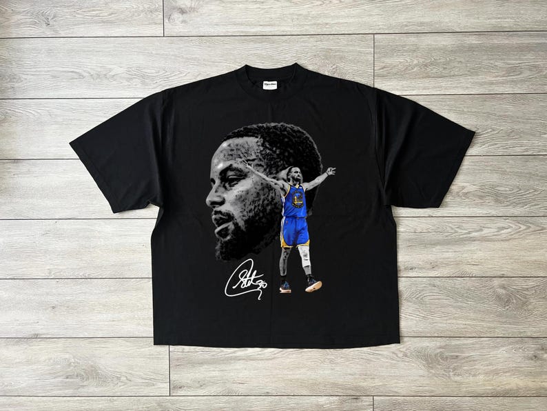 May include: Black t-shirt featuring a grayscale portrait and a full-color image of a basketball player in a blue and gold uniform. The shirt also includes a signature in white.