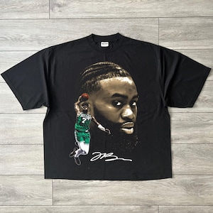 Puede incluir: Camiseta negra con un retrato detallado de un jugador de baloncesto, y una imagen más pequeña de él en acción, vistiendo una camiseta verde y blanca. La camiseta también incluye la firma del jugador.