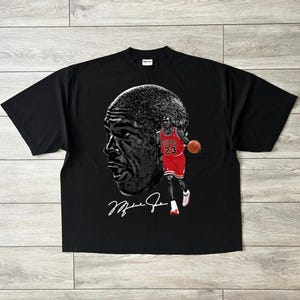 May include: Black t-shirt featuring a large grayscale image of a basketball player's face and a full-body image of the player in a red and black uniform. The player is holding a basketball. The shirt also has the player's signature.
