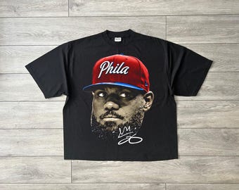May include: A black short-sleeve t-shirt featuring a graphic print of a person wearing a red and blue baseball cap with the word Phila written in white script. Below the face is a white stylized signature and the number 23 in white ink.