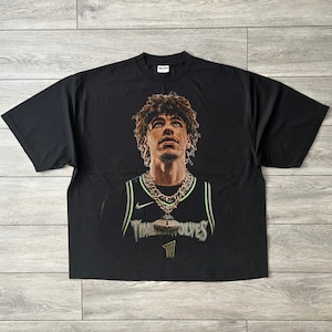 May include: A black short-sleeve t-shirt featuring a graphic print of a basketball player with curly hair wearing a jersey with the text TIMBERWOLVES and the number 1. The player is depicted wearing a thick gold chain with a large pendant.