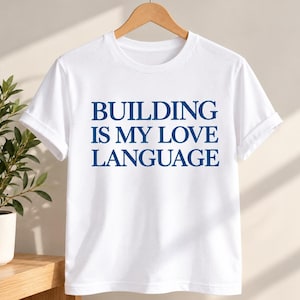 Peut inclure: Un t-shirt blanc à manches courtes avec le texte bleu foncé "BUILDING IS MY LOVE LANGUAGE" imprimé dans une police serif grasse au centre de la poitrine. Le vêtement présente un col rond classique et des manches à revers, sur un cintre.