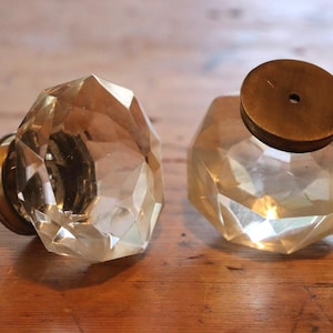 May include: Two faceted crystal cabinet knobs with brass fittings. The knobs have a geometric design and a warm, amber colour. One knob is attached, and the other is separate, showing the brass base. Suitable for furniture restoration projects.