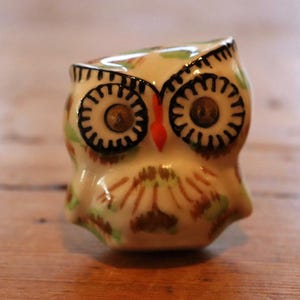 May include: A ceramic owl ornament with a cream base, featuring black, brown, green, and orange detailing. The owl has large, circular eyes and a small, pointed beak. This figurine is a decorative item.