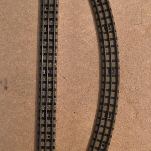 May include: Two sections of model railway track. One is straight, the other is curved. Both are black with a tan and green ballast. The tracks are made of plastic and are designed for a model railway set.