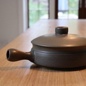 May include: A grey ceramic casserole dish with a lid and a handle. The dish has a round body with horizontal ridges and a dark brown handle. The lid has a small, round knob. The dish is on a light brown wooden surface.