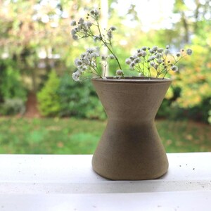 May include: A small, brown ceramic vase, shaped like an hourglass, contains delicate white flowers. The vase is on a white surface, with a blurred green and brown backdrop of trees and foliage.