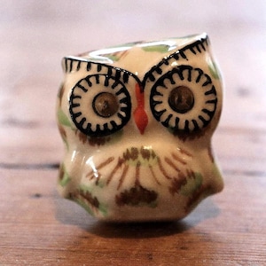 May include: A small ceramic owl figurine featuring a cream-coloured base with hand-painted details in brown and light green. The owl has large, circular eyes outlined in black, a small orange beak, and stylised feather markings across its body.